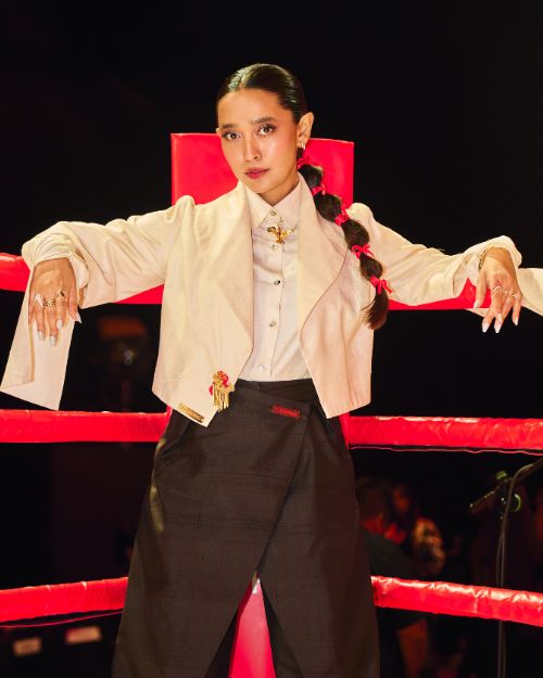 Woman leaning on red boxing ring ropes, wearing a cream blazer and white shirt, with a long braided ponytail.